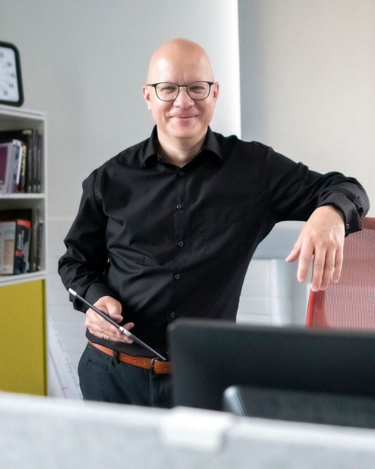 Christian Rahn, Marketing-Experte, steht lächelnd in einem modernen Büro. Er trägt ein schwarzes Hemd, eine Brille und hält ein Tablet in der Hand. Mit dem rechten Arm lehnt er locker an einem roten Bürostuhl. Im Hintergrund sind Bücherregale und ein Wanduhr-Ausschnitt zu sehen.