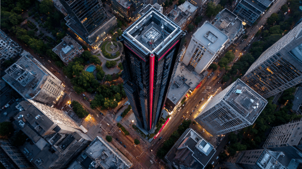 Drohnenaufnahme eines modernen Wolkenkratzers inmitten einer dicht bebauten Innenstadt bei Dämmerung. Das Hochhaus mit markanter vertikaler Beleuchtung (rot) steht im Zentrum des Bildes und dominiert die Szenerie. Umgeben von weiteren Hochhäusern, Straßen mit fließendem Verkehr und grünen Stadtparks vermittelt das Bild den Eindruck von urbaner Dichte, Dynamik und vertikaler Dominanz – ein Symbol für strategische Positionierung, Sichtbarkeit und Standfestigkeit inmitten komplexer Umfelder.